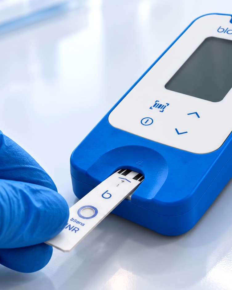 A blue-gloved hand inserting a bSens.INR strip into the bSens One reader.